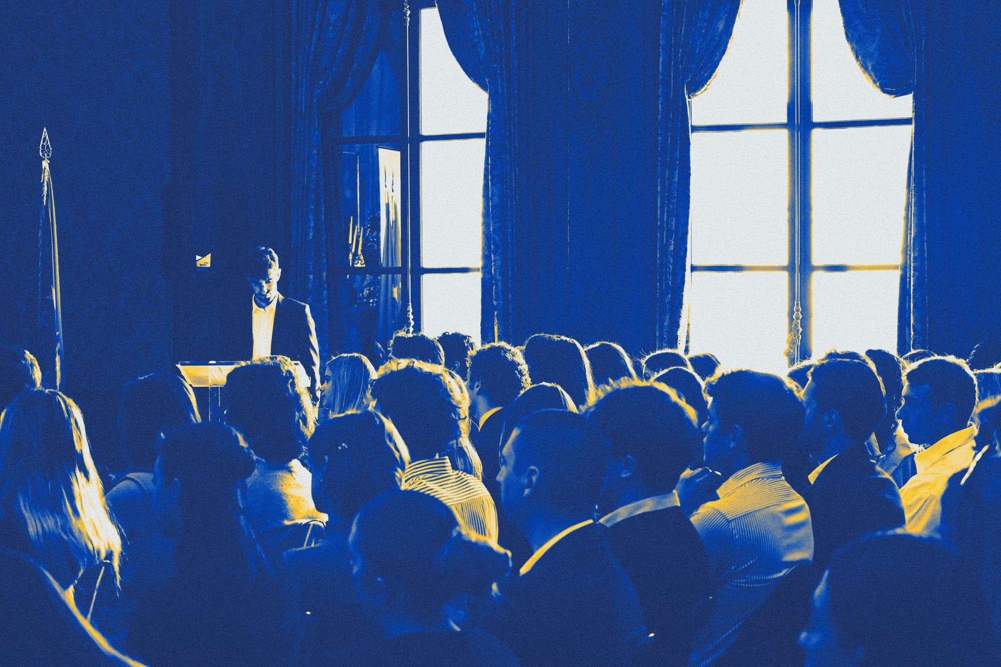 An audience facing tall curtained windows, blue and yellow duotone