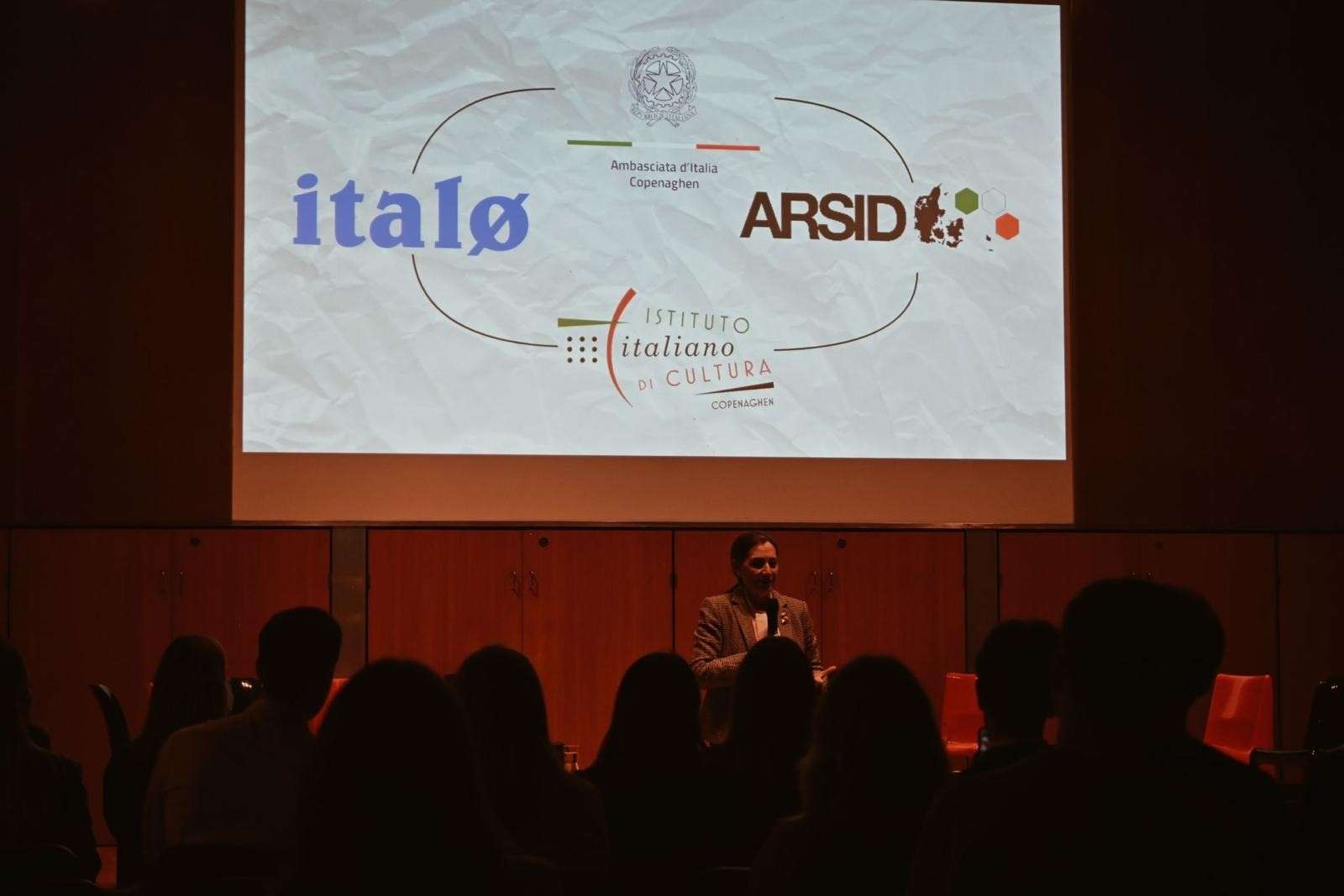 Talk in a dark auditorium, the screen showing the italø, ARSID and Istituto Italiano di Cultura logos