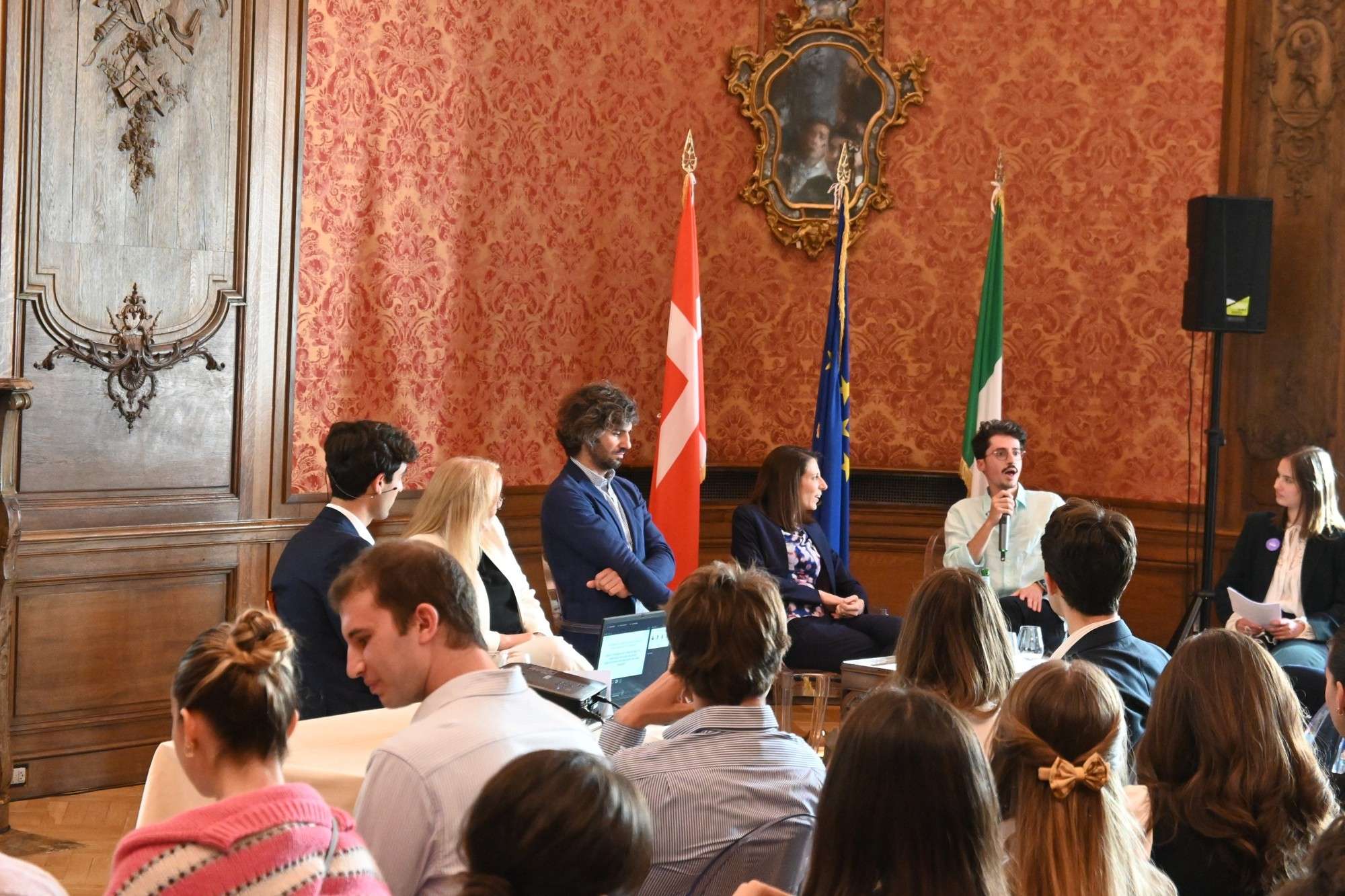 Speakers on a panel in a red damask room with Italian, EU and Danish flags, audience in the foreground