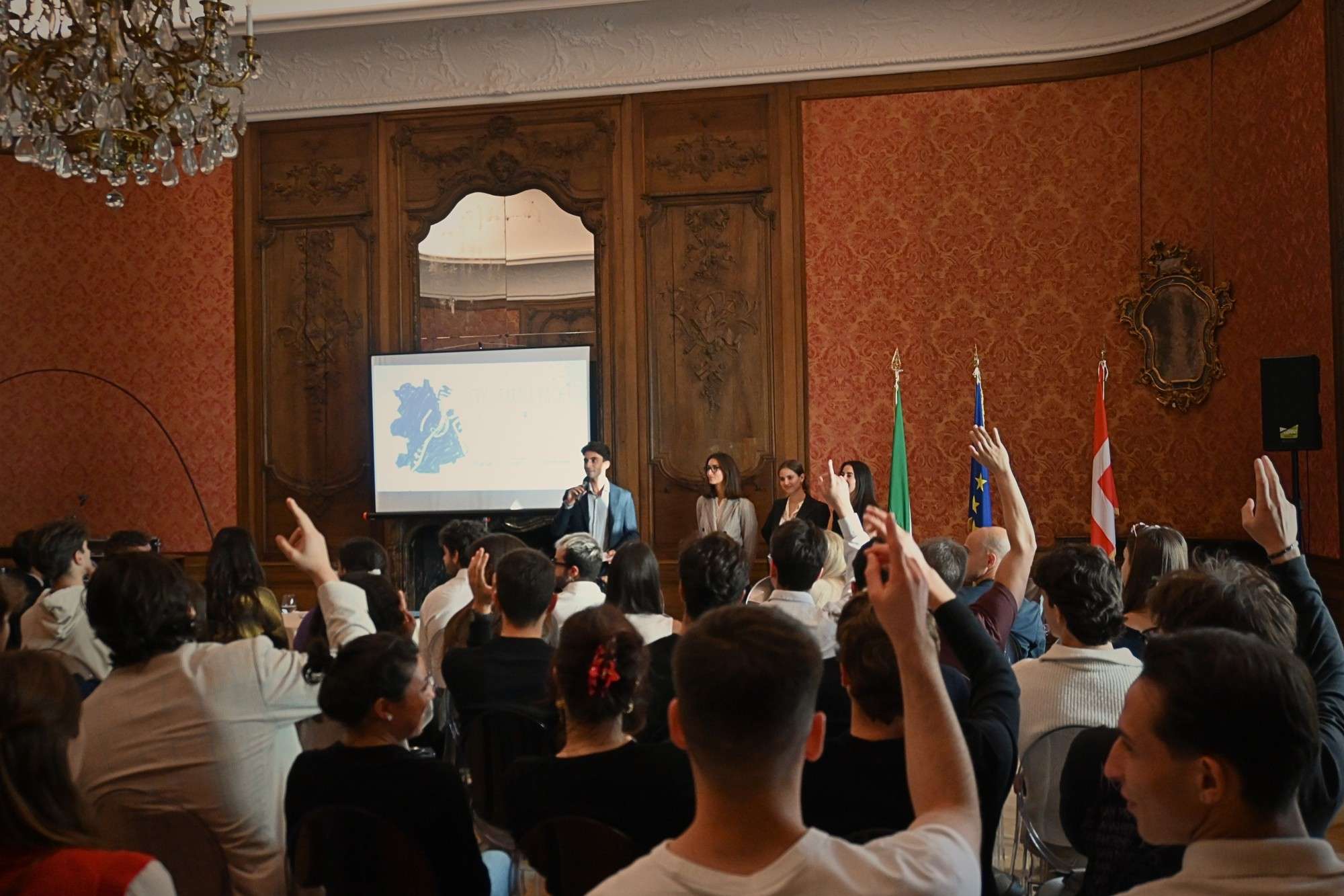 A packed room raising hands during an italø talk, with Italian, Danish and EU flags beside the speakers
